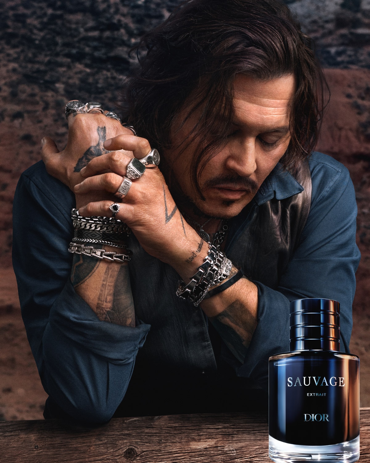 Johnny Depp se sklopenou hlavou a rukama sepjatýma před sebou, na sobě má tmavě modrou košili a koženou vestu, s mnoha prsteny a náramky, s lahvičkou parfému Dior Sauvage Extrait v popředí.