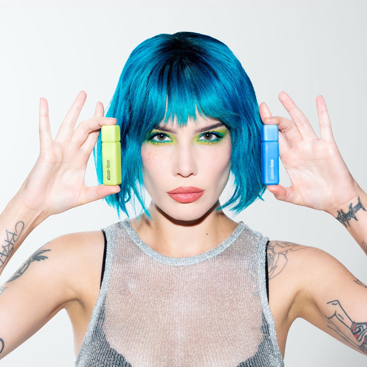 Žena s modrými vlasy drží v každé ruce barevný flakonek: v jedné ruce světle zelený a v druhé modrý.