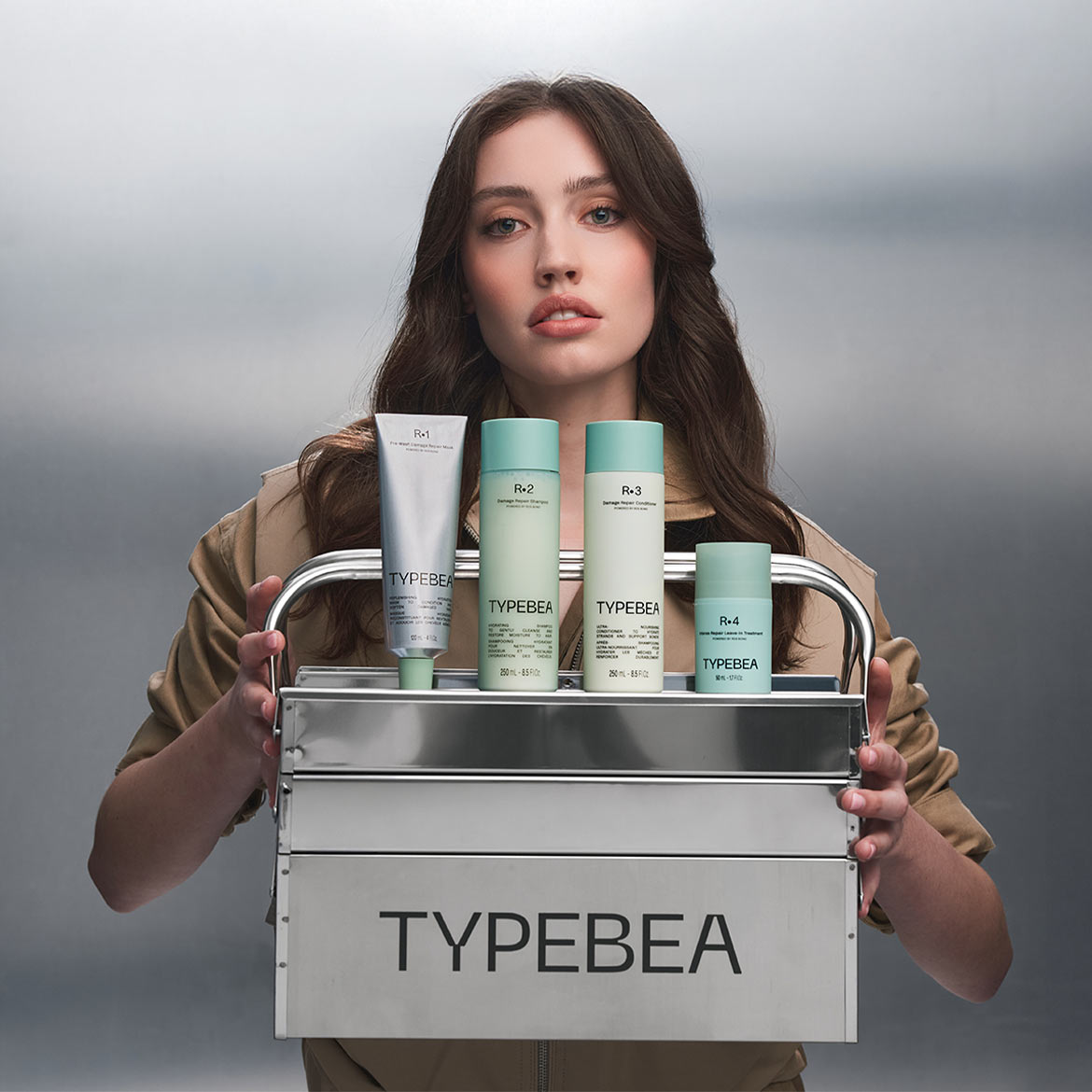 Žena drží stříbrný box se čtyřmi produkty péče o vlasy Typebea, včetně šamponu, kondicionéru a masky, vše v bílých lahvích s mátově zelenými víčky.