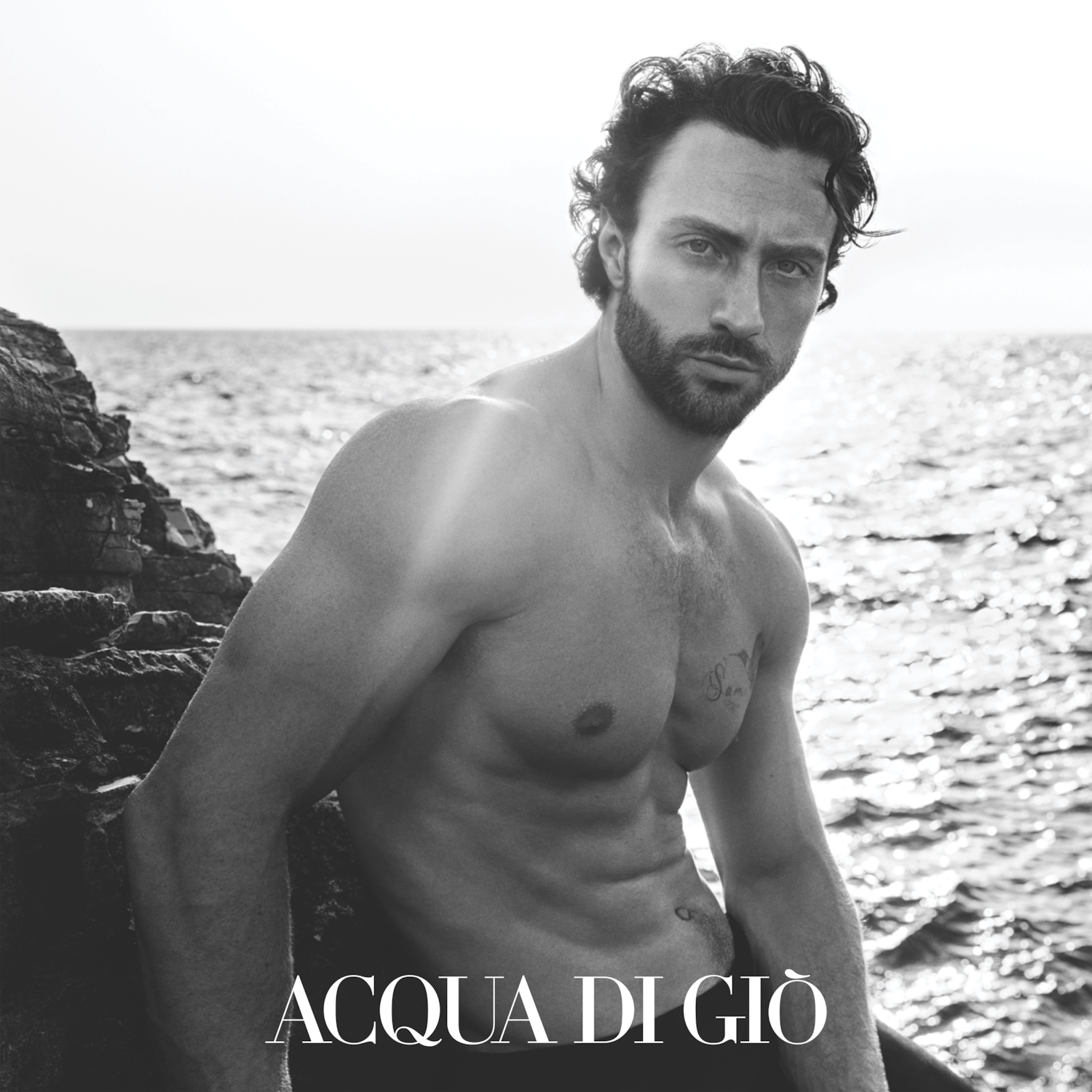 Černobílý snímek muže s vlnitými vlasy a vousy, který sedí na skalnatém pobřeží s nápisem ACQUA DI GIÒ.