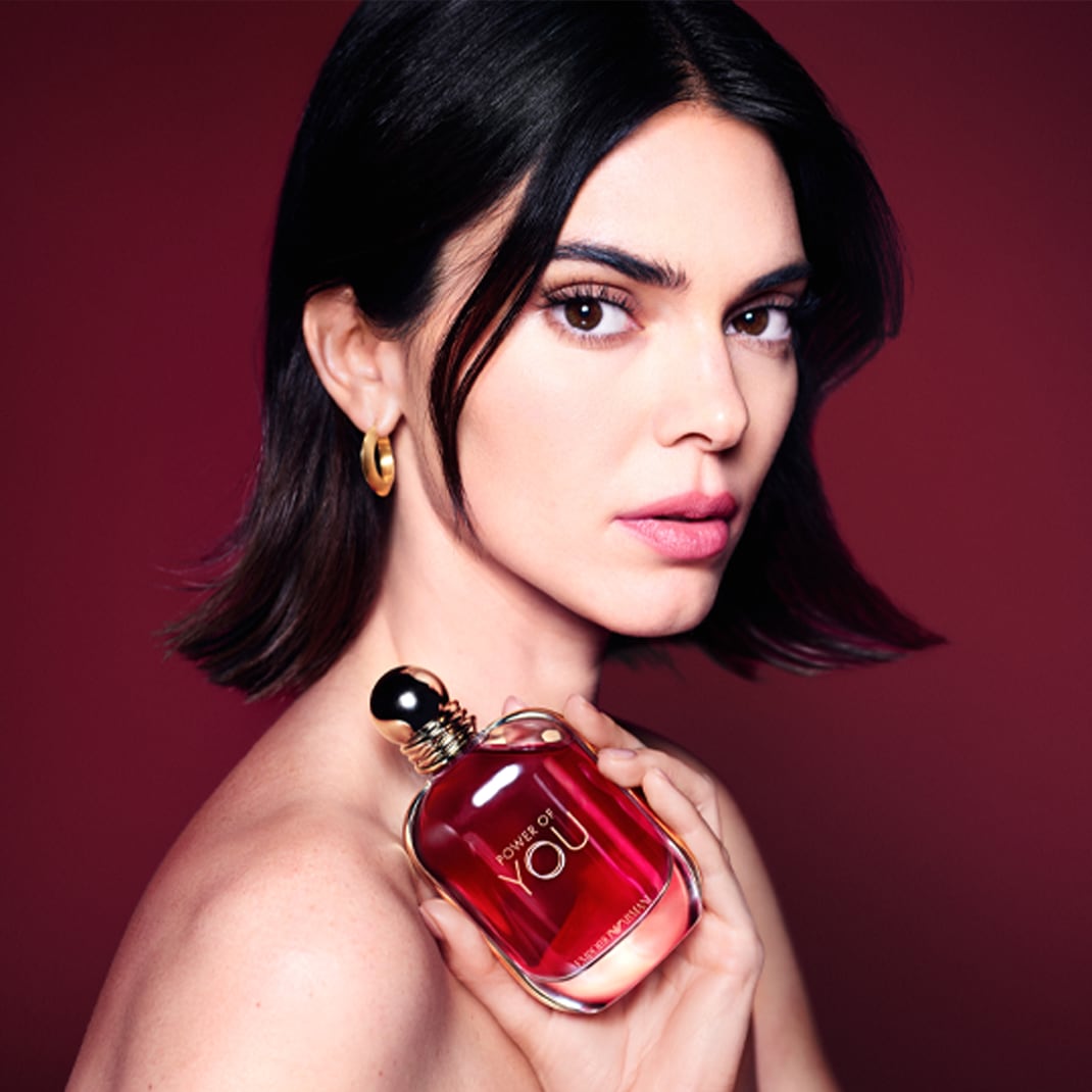Portrét ženy s krátkými tmavými vlasy a zlatými náušnicemi, která drží v ruce flakon parfému Power of You od Armaniho v červeno-zlatém provedení.