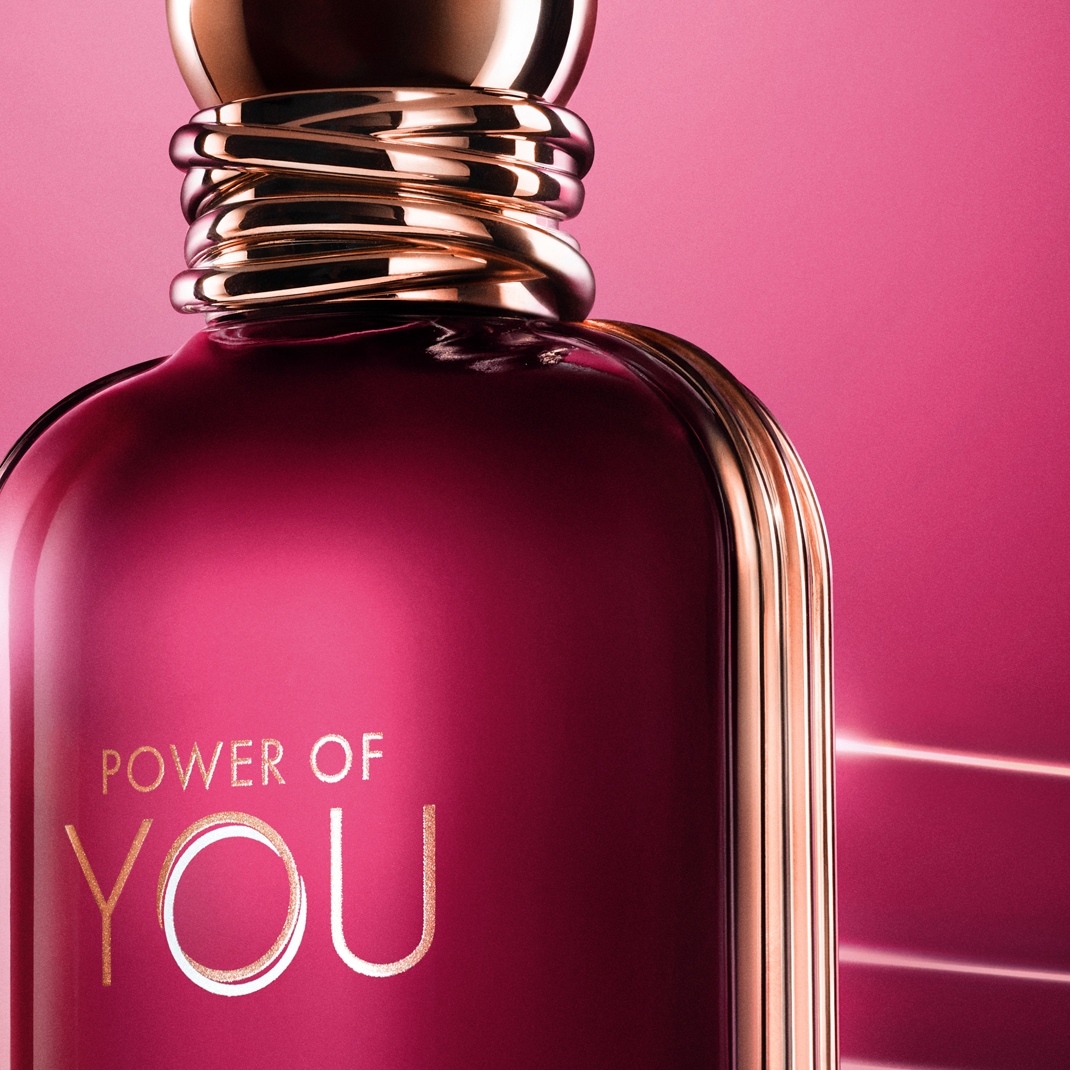 Detailní záběr na flakon parfému s nápisem „POWER OF YOU“ na sytě růžovém pozadí, doplněný o zlaté akcenty na hrdle a okrajích.