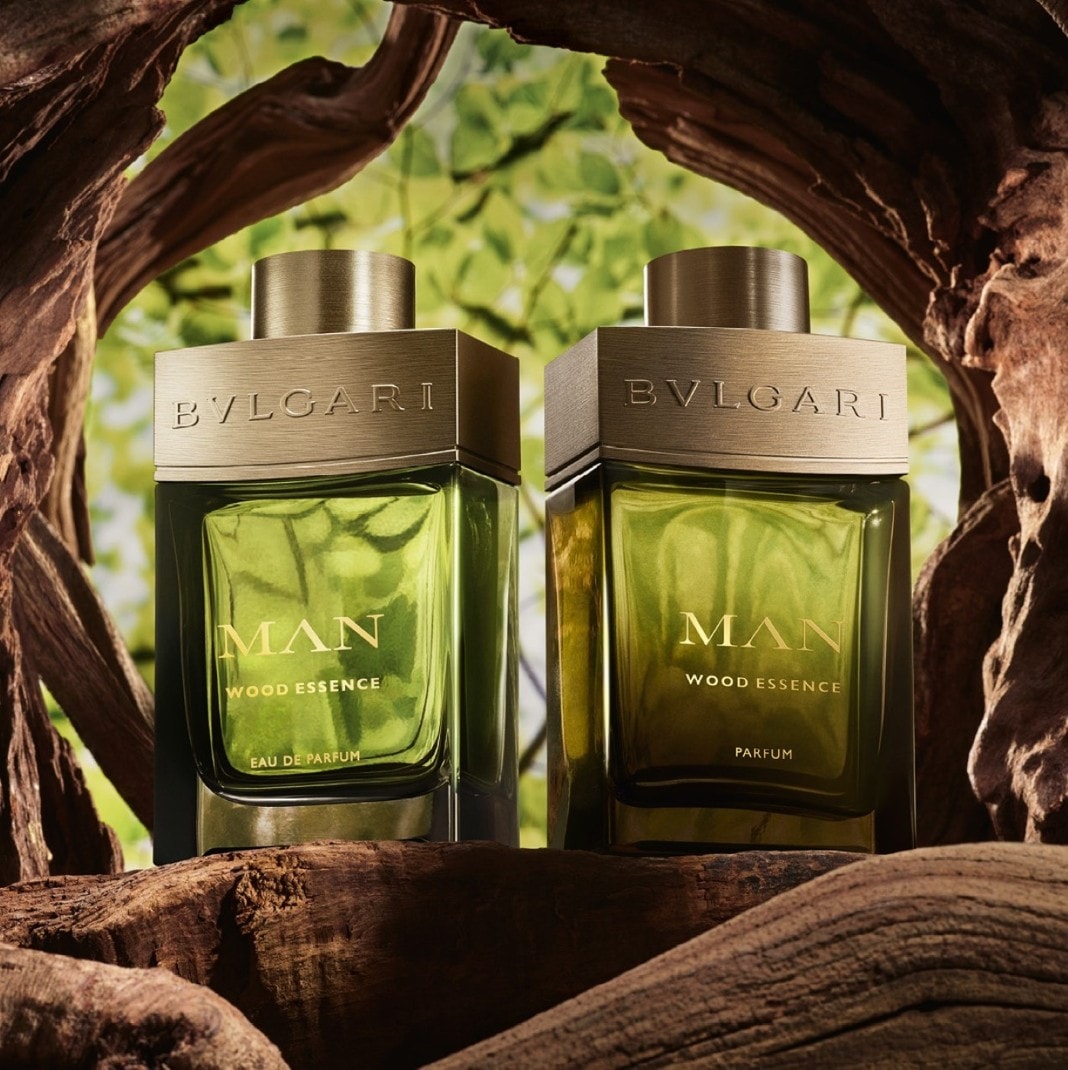 Dva flakony pánské vůně Bvlgari Man Wood Essence s dřevitým aroma, umístěné na kusu dřeva s pozadím zelených listů.