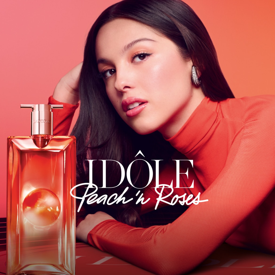 Na oranžovém pozadí leží Olivia Rodrigo vedle flakonu parfému Idôle Peach 'n Roses od Lancôme.