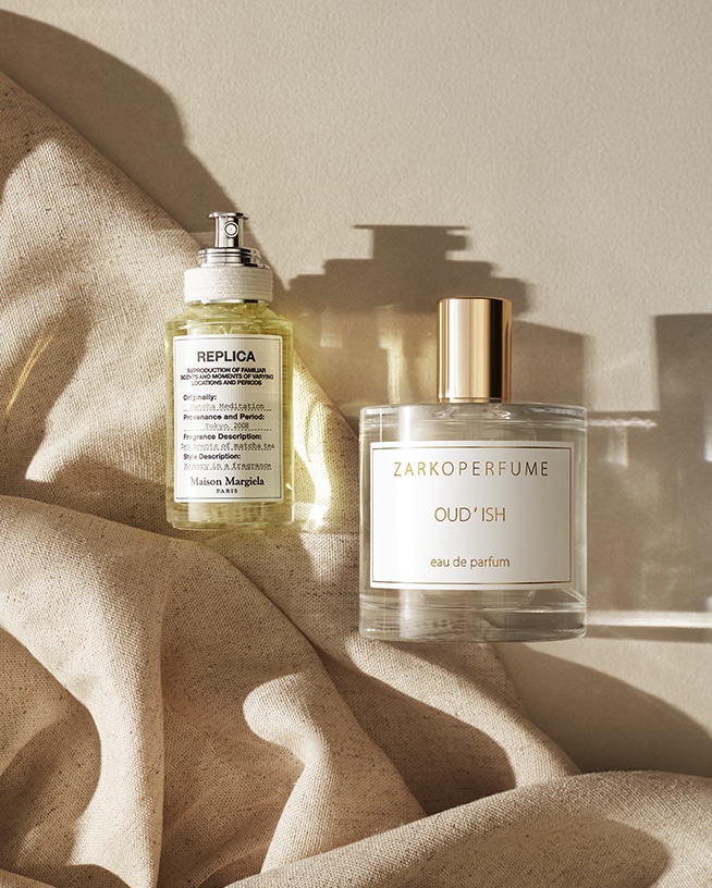 Dva flakony parfémů, jeden od Maison Margiela s etiketou REPLICA a druhý od ZARKOPERFUME s názvem OUD'ISH, stojí na béžové látce.