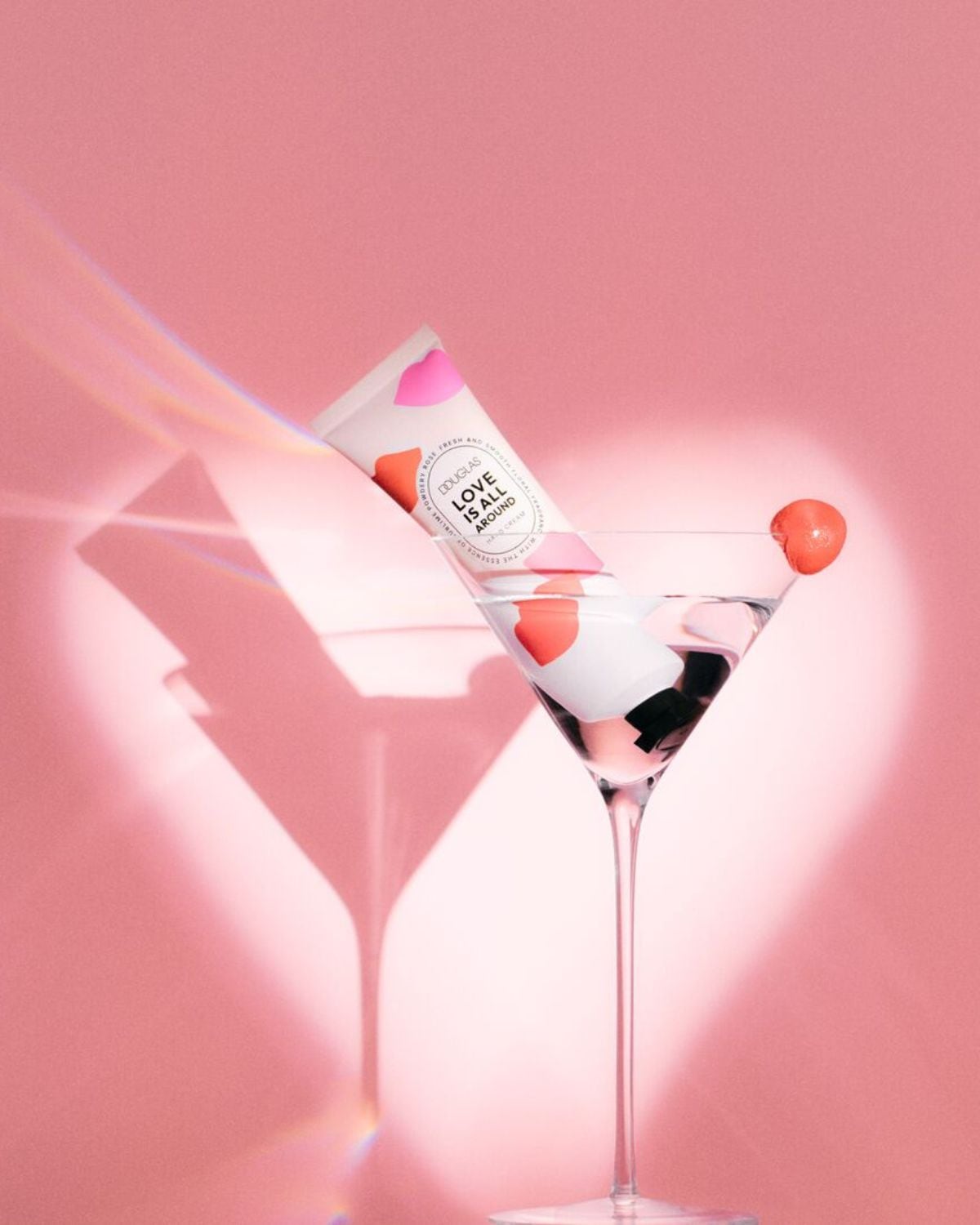 Růžové pozadí s koktejlovou sklenicí, v níž je tuba krému na ruce Douglas Love Is All Around s růžovými a červenými srdíčky.