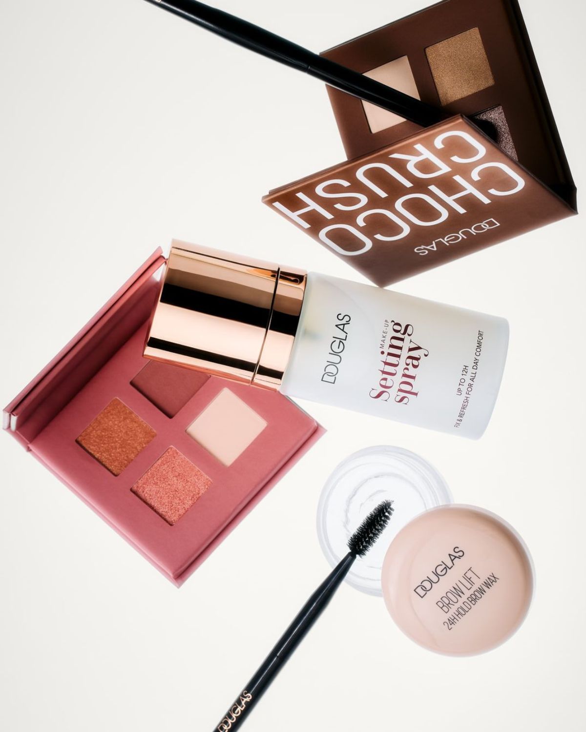 Dekorativní kosmetika Douglas zahrnuje oční stíny Choco Crush v hnědé paletce, růžovou paletku očních stínů, fixační sprej s růžovo-zlatým víčkem a vosk na obočí Brow Lift s černým kartáčkem.