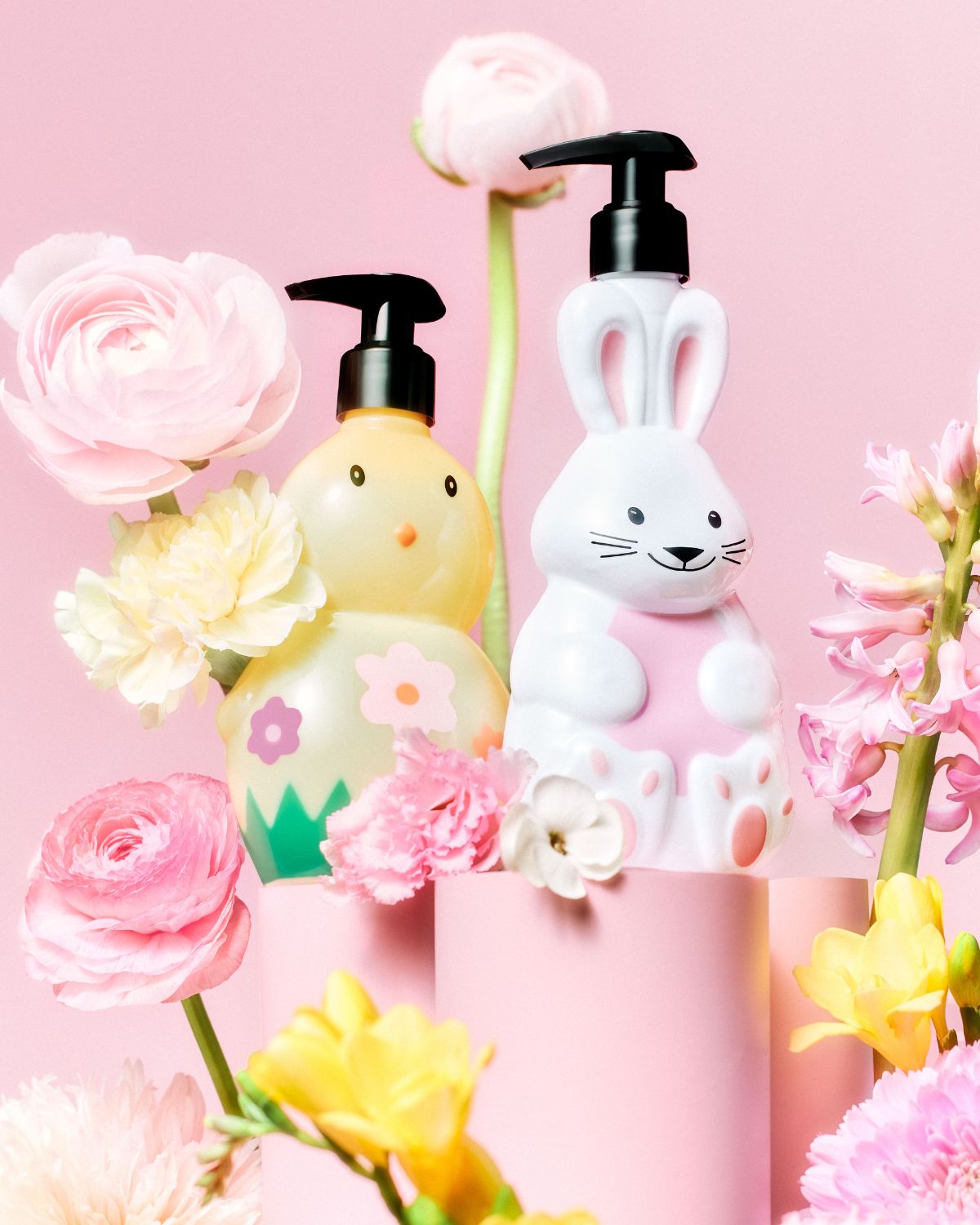 Na růžovém pozadí stojí žlutá láhev ve tvaru kuřátka a bílá láhev ve tvaru králíčka s černými pumpičkami, obklopené růžovými a žlutými květy.