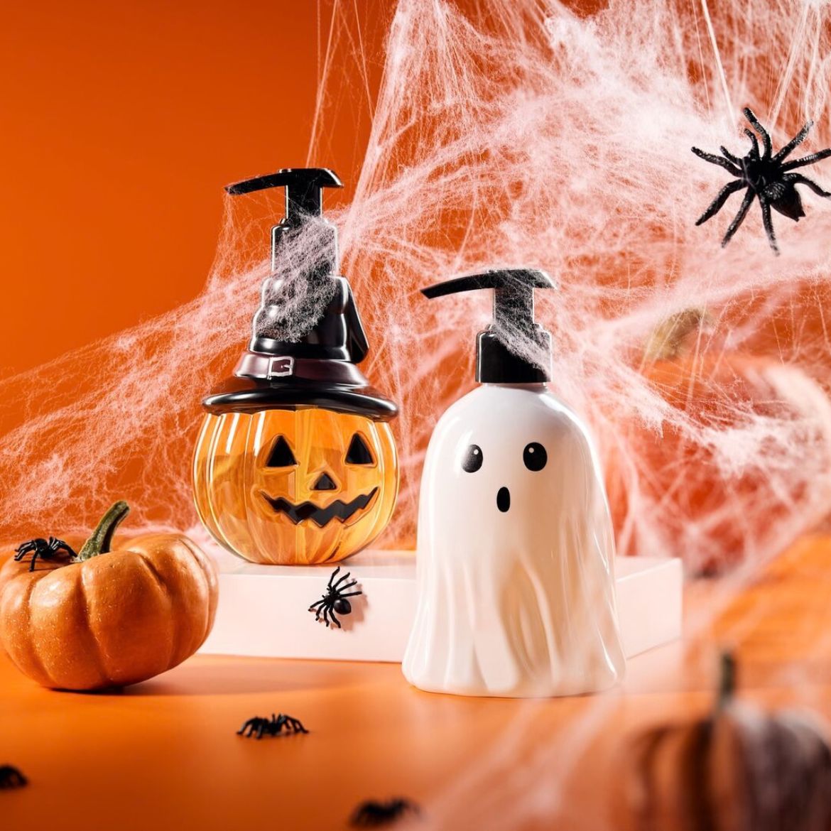 Dvě halloweenské lahvičky s pumpičkou, jedna ve tvaru dýně s černým kloboukem a druhá jako bílý duch, stojí na oranžovém pozadí s pavučinami a plastovými pavouky.