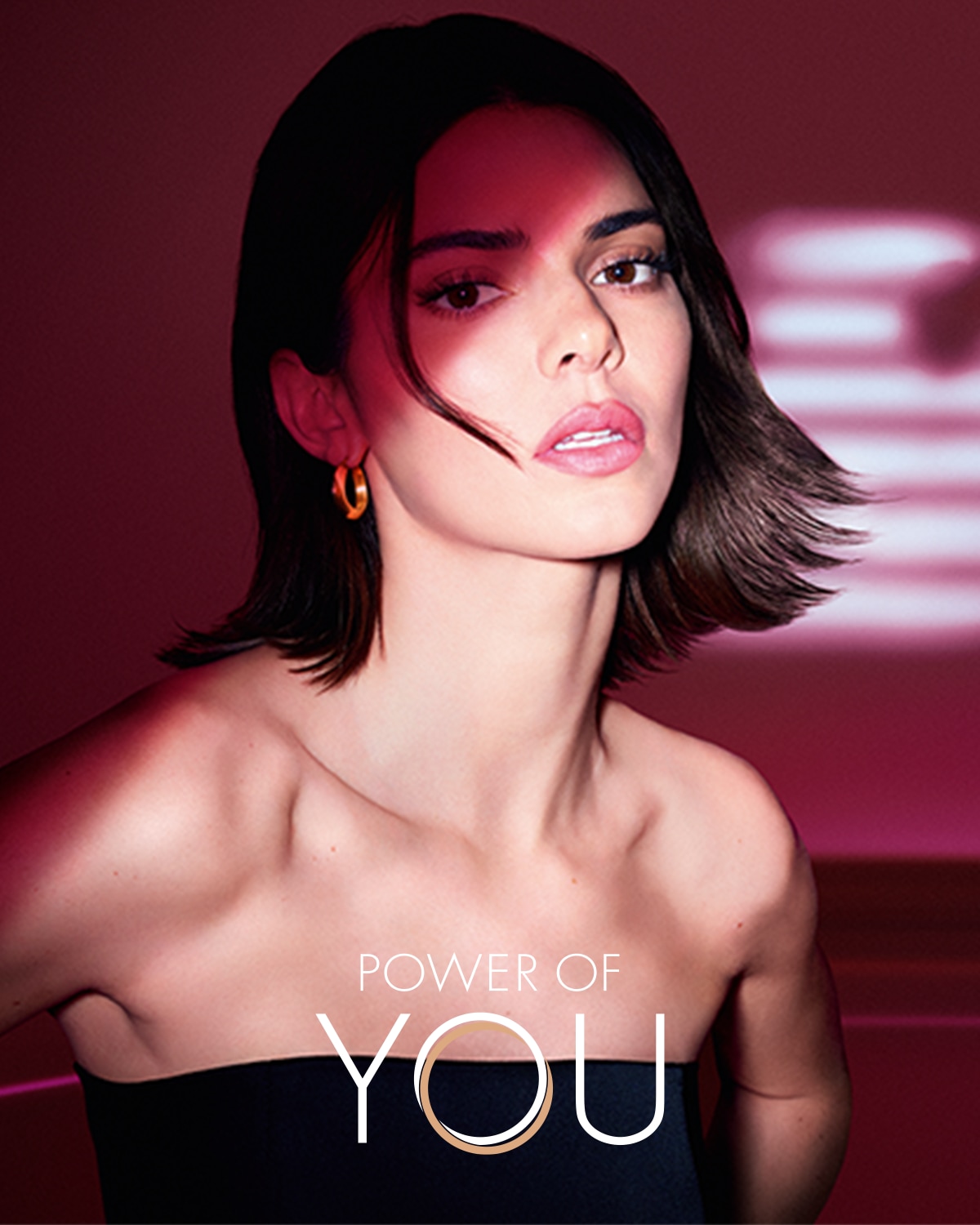 Na portrétu je žena s krátkými hnědými vlasy, která má na sobě černé šaty bez ramínek a zlaté náušnice, v pozadí je růžové světlo a nápis "POWER OF YOU".