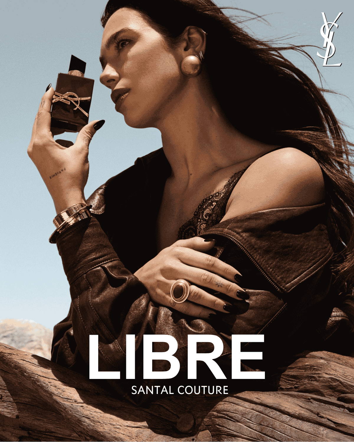 Žena s dlouhými tmavými vlasy drží v ruce parfém Libre Santal Couture od Yves Saint Laurent, který má tmavou lahvičku se zlatým logem YSL a černým uzávěrem, na sobě má hnědou koženou bundu a krajkový top.